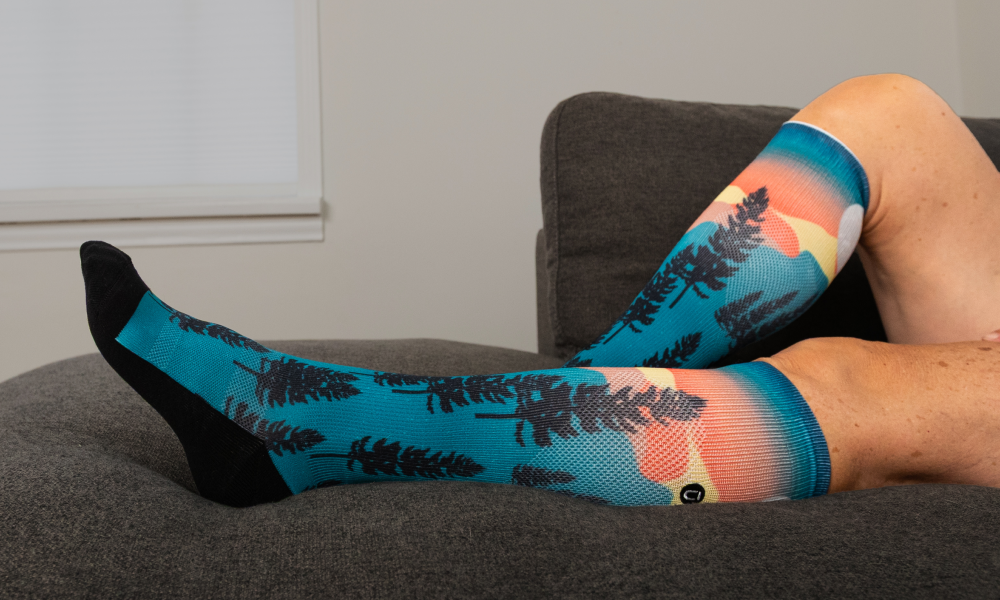 Person resting on a couch wearing OUTWAY teal knee-high compression socks with tree and sunset pattern
