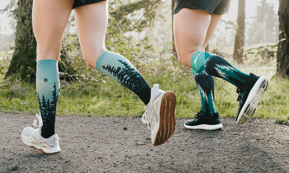 Two runners on an outdoor trail wearing OUTWAY knee-high compression socks with a teal forest tree design, shot from behind at ground level showing legs mid-stride.