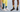 Two people wearing colourful OUTWAY crew socks, one in a teal mountain forest design with white sneakers and one in a yellow sunset landscape design with black sneakers, shown from the waist down against a light blue background