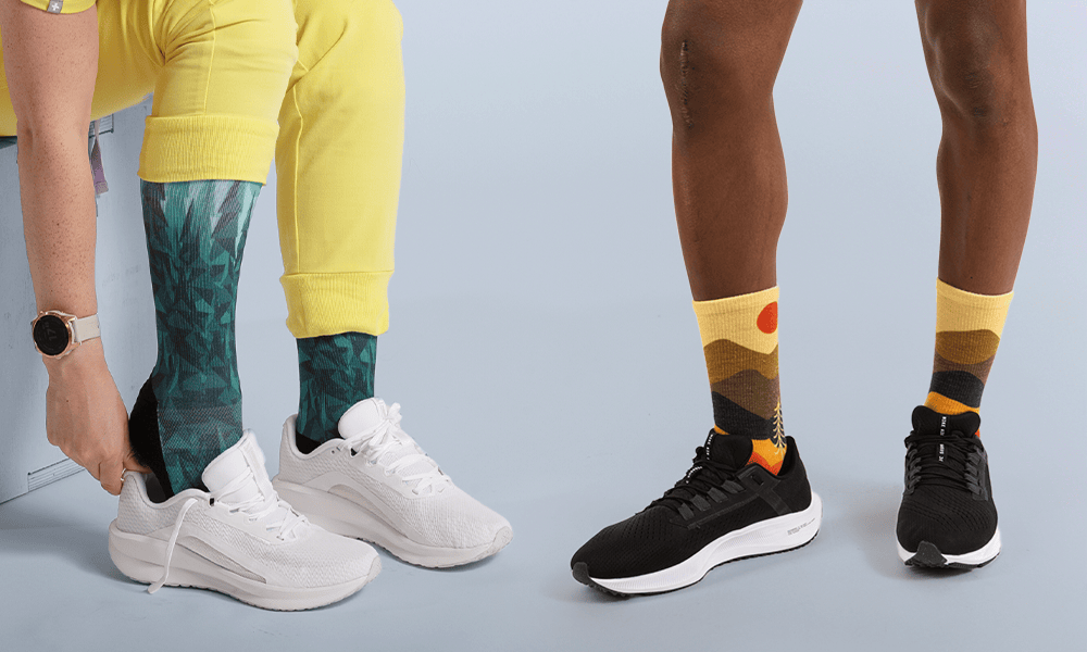 Two people wearing colourful OUTWAY crew socks, one in a teal mountain forest design with white sneakers and one in a yellow sunset landscape design with black sneakers, shown from the waist down against a light blue background