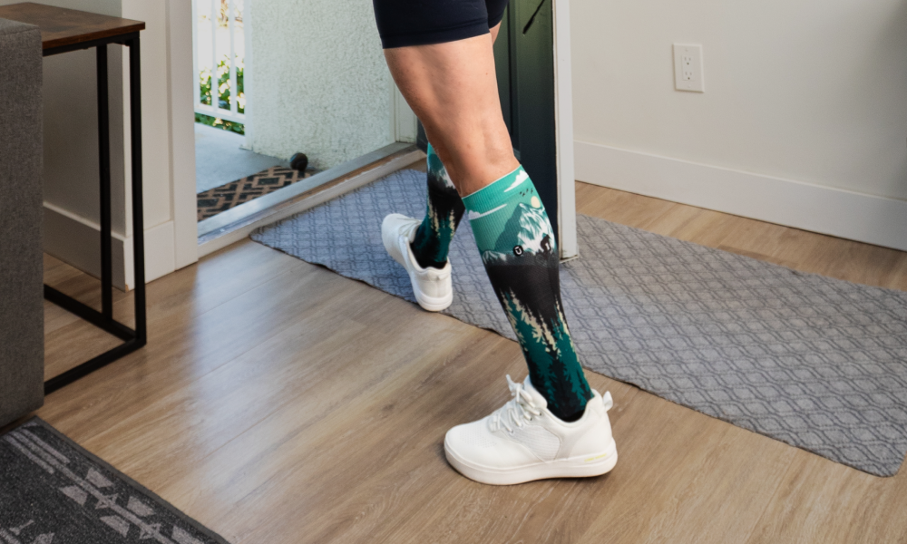 Person walking indoors wearing OUTWAY knee high compression socks with green and black mountain pattern