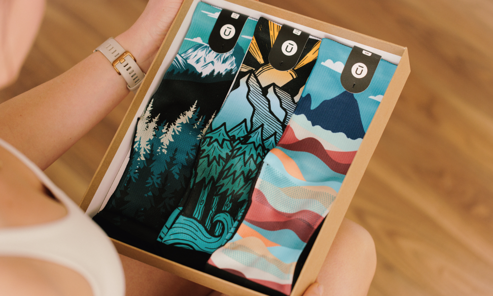 Person holding an OUTWAY sock 3-pack gift box containing three pairs of athletic crew socks with nature-inspired designs